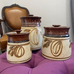 May include: Set of three ceramic canisters with lids. The canisters are brown and white with a speckled glaze and a leaf and oval design. The lids are brown.