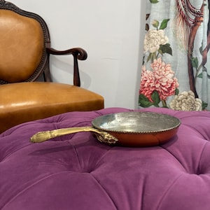 May include: A round copper pan with a silver-toned interior and a decorative brass handle. The exterior of the pan features a smooth copper finish, while the handle is detailed with ornate metalwork where it attaches to the side of the pan.