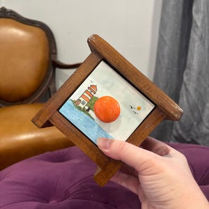 May include: Wooden box with a ceramic tile featuring a lighthouse scene. The tile has a white background with a lighthouse, blue water, and an orange sun. The box has a round orange knob.