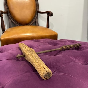 May include: An antique auger drill with a wooden handle and a metal drill bit, resting on a tufted purple ottoman. A vintage leather chair is in the background.