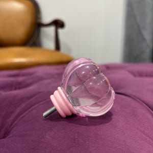 May include: A clear, heart-shaped cabinet knob with a pink base and screw. The knob is made of glass and has a faceted design. The knob is resting on a purple velvet surface.