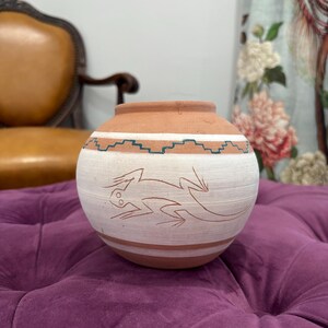 May include: A round terracotta pottery vessel featuring a wide white band around the center. The white section displays a hand-etched lizard illustration, while a teal-colored geometric zigzag pattern encircles the upper portion of the pot.