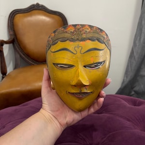 May include: A yellow wooden mask with a serene expression. The mask has detailed features, including closed eyes, a small smile, and intricate headwear with orange and gold accents. The mask is held in a hand against a purple tufted surface.
