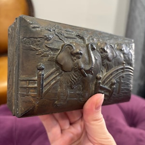 May include: A rectangular bronze-colored box with a detailed relief of elephants, trees, and a fence. The box has a slightly aged appearance, with visible wear on the edges. The relief depicts multiple elephants in a naturalistic setting.
