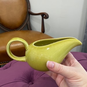 May include: A green ceramic gravy boat with a rounded body and a curved spout. The boat has a smooth, glossy finish.