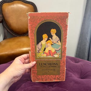 May include: Vintage cookbook titled "The Calendar of Luncheons Teas and Suppers" by Elizabeth Hiller. The cover is a faded red with an illustration of three women at a table. The book has a gold title and is held by a person.