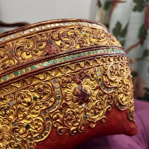 May include: Ornate, gold-colored decorative object with intricate carvings and embedded jewels. The object has a red base and is adorned with detailed patterns and embellishments. The design suggests a cultural or historical artifact.