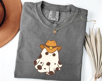 May include: A gray Comfort Colors t-shirt featuring a cartoon ghost wearing a cowboy hat with a gold star. The ghost has brown spots. A silver necklace with a small pendant is on the shirt. A brown cowboy hat is in the background.