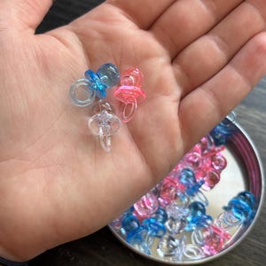 May include: A hand holds three small, clear plastic pacifiers in blue, pink, and clear. A small, silver container holds many more pacifiers in the same colors. The pacifiers are a baby-themed item.