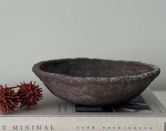 May include: A dark brown, textured bowl with a rough, uneven rim sits atop a book. Beside the bowl are two sprigs of reddish-brown decorative flowers. The book's cover reads "SOFT MINIMAL".