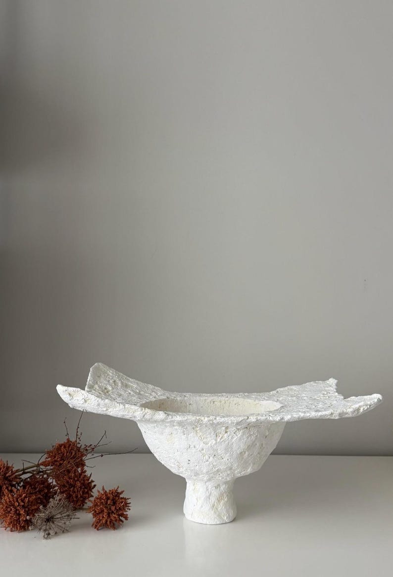 May include: A textured white ceramic bowl with a wide, irregular rim and a pedestal base. The bowl has a rough, handmade appearance and is set against a neutral background. Dried, reddish-brown floral elements are visible on the left.