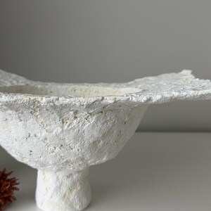 May include: A textured, white, handmade bowl with a wide, irregular rim. The bowl has a rounded body and sits on a short, pedestal base. The surface has a rough, organic appearance, suggesting a natural material. The bowl is a decorative item.