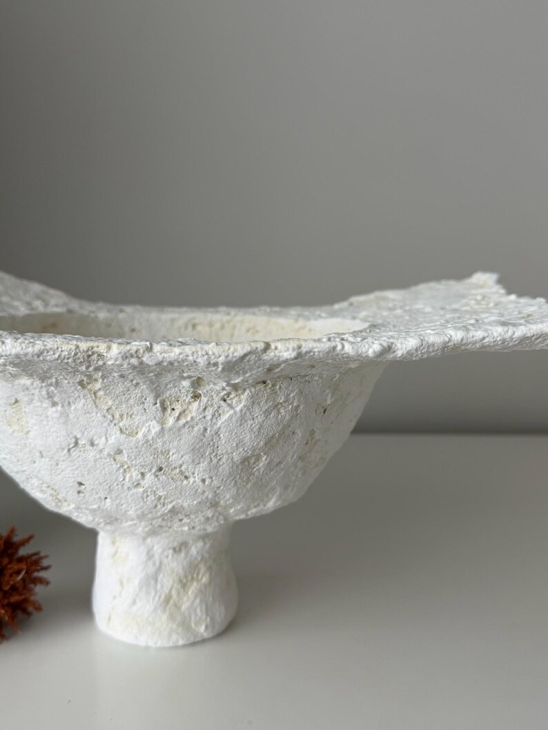 May include: A textured, white, handmade bowl with a wide, irregular rim. The bowl has a rounded body and sits on a short, pedestal base. The surface has a rough, organic appearance, suggesting a natural material. The bowl is a decorative item.