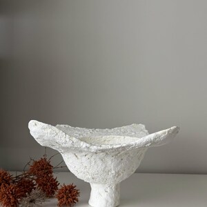 May include: A white ceramic mug with a black handle and the text "But First, Coffee" in a bold, black font. The mug is set against a neutral background, highlighting its simple design.