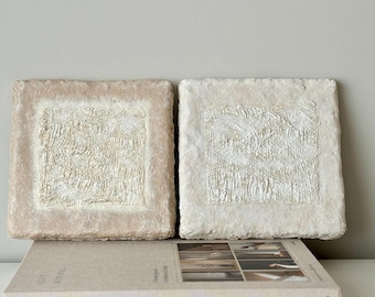 May include: Two square-shaped wall art pieces with a textured white design on a beige background. The pieces are made of paper and have a rough, handmade look.