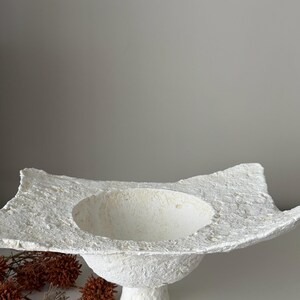 May include: A textured, white, handmade bowl with a wide, irregular rim and a rounded base. The bowl has a central, circular depression and sits on a short, pedestal-style base. Decorative dried flowers are in the background.