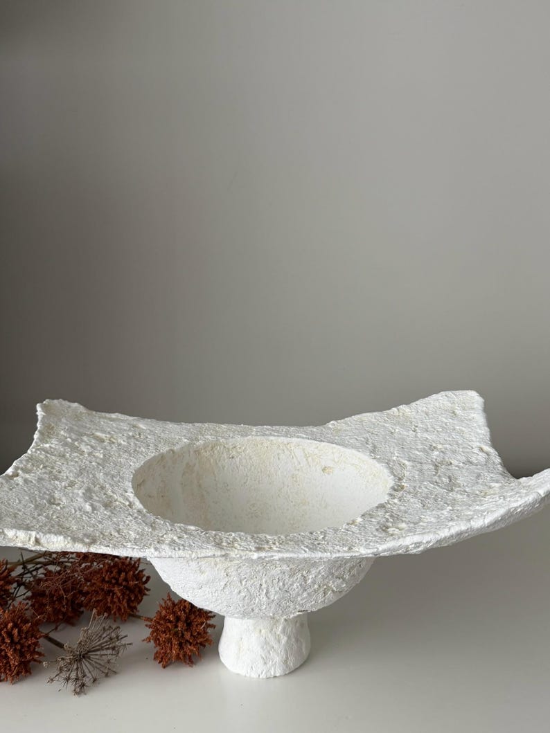 May include: A textured, white, handmade bowl with a wide, irregular rim and a rounded base. The bowl has a central, circular depression and sits on a short, pedestal-style base. Decorative dried flowers are in the background.