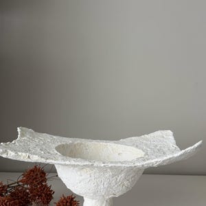 May include: A white, textured, paper-like bowl with a wide, irregular rim. The bowl sits on a pedestal base. Dried, brown floral elements are arranged to the left of the bowl. The bowl is a decorative item.