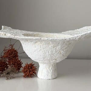 May include: A textured white decorative bowl with a wide, irregular rim, set on a pedestal base. The bowl has a rough, handmade appearance. Dried, reddish-brown floral elements are arranged to the left of the bowl.