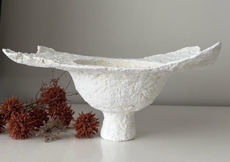 May include: A textured white decorative bowl with a wide, irregular rim, set on a pedestal base. The bowl has a rough, handmade appearance. Dried, reddish-brown floral elements are arranged to the left of the bowl.