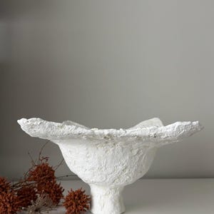 May include: A textured white decorative bowl with a pedestal base and a ruffled rim. The bowl has a rough, handmade appearance and is set against a neutral background. Dried orange flowers are in the foreground.