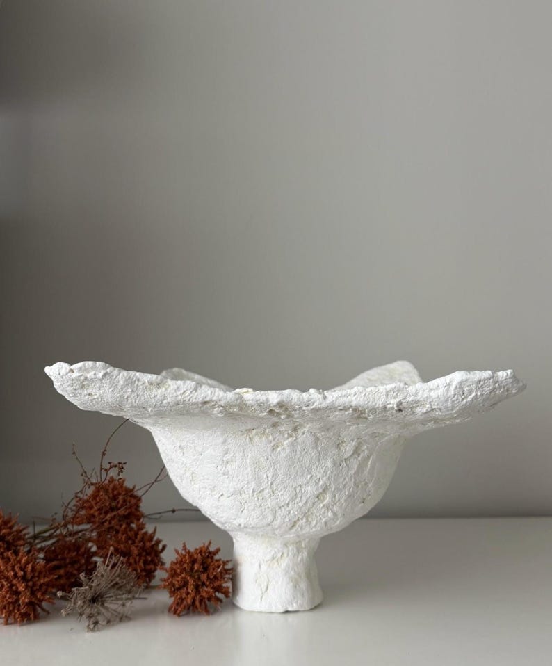 May include: A textured white decorative bowl with a pedestal base and a ruffled rim. The bowl has a rough, handmade appearance and is set against a neutral background. Dried orange flowers are in the foreground.