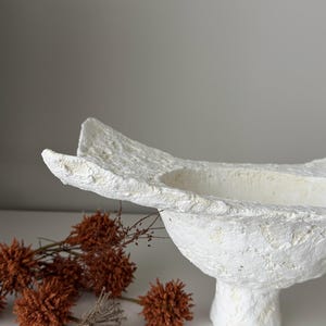 May include: A textured white decorative bowl with a pedestal base. The bowl has an organic shape with a wide opening. Dried orange flowers are arranged near the bowl, adding a natural element to the composition.