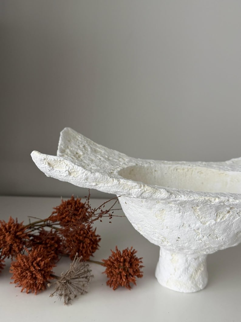 May include: A textured white decorative bowl with a pedestal base. The bowl has an organic shape with a wide opening. Dried orange flowers are arranged near the bowl, adding a natural element to the composition.