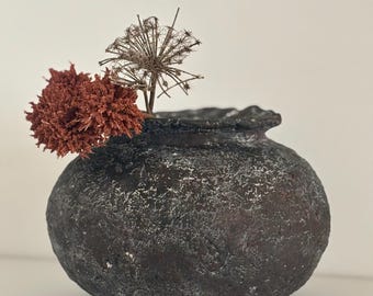 May include: A dark gray, textured ceramic vase with a rounded shape, holding dried flowers. The vase has a rough, uneven surface and a slightly uneven rim.