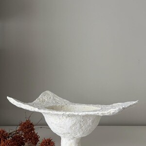 May include: A textured white paper mache bowl with a wide, undulating rim. The bowl sits on a pedestal base. Dried, reddish-brown floral elements are visible to the left of the bowl. The bowl's design is organic and sculptural.