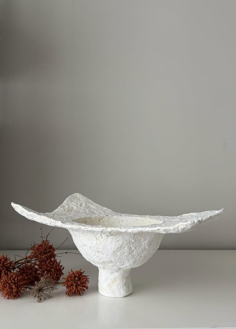 May include: A textured white paper mache bowl with a wide, undulating rim. The bowl sits on a pedestal base. Dried, reddish-brown floral elements are visible to the left of the bowl. The bowl's design is organic and sculptural.