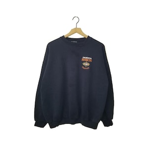 May include: A dark navy blue crewneck sweatshirt featuring long sleeves and ribbed cuffs. On the upper left chest, there is an embroidered graphic with the text "DENVER BRONCOS" in white and orange, along with a small logo and the word "CHAMPIONS".