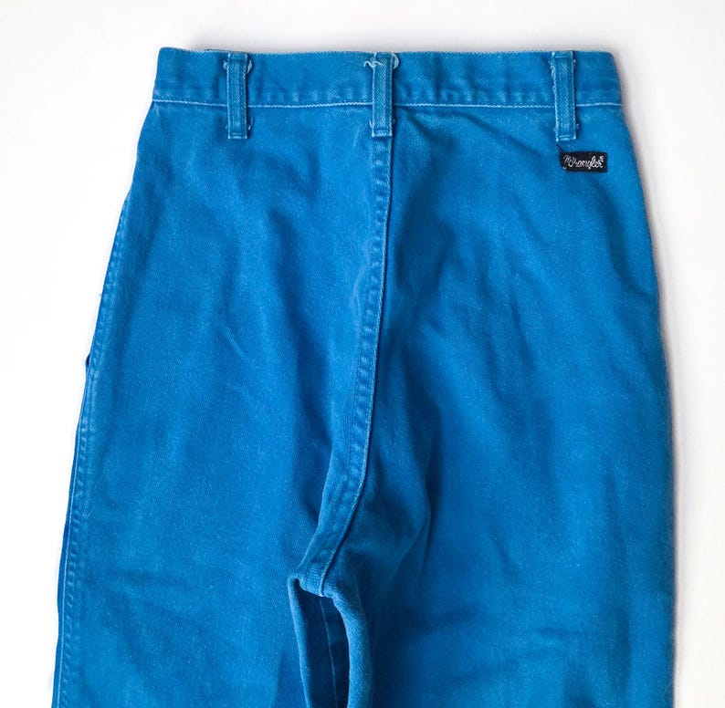 May include: Close-up of bright blue denim pants. The pants have a classic design with belt loops, a back yoke, and a small black and white label. The stitching is white, providing contrast against the blue fabric.