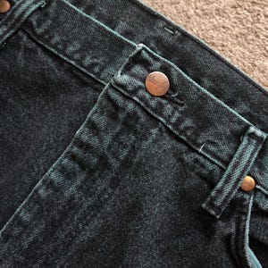 以下が含まれることがあります： 濃いティールデニムジーンズのクローズアップ。ジーンズは、ブランド名「Wrangler」とマッチするリベットが付いた銅色のボタンが特徴です。生地は、ステッチとベルトループが見える、テクスチャード加工された外観をしています。