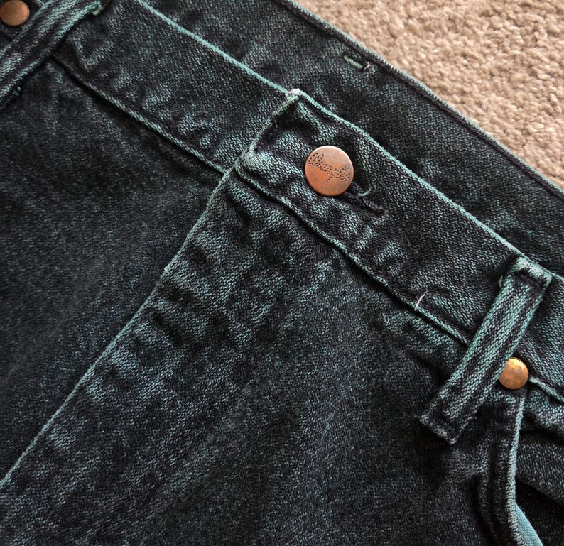 以下が含まれることがあります： 濃いティールデニムジーンズのクローズアップ。ジーンズは、ブランド名「Wrangler」とマッチするリベットが付いた銅色のボタンが特徴です。生地は、ステッチとベルトループが見える、テクスチャード加工された外観をしています。