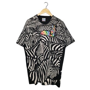 May include: Black and white zebra print t-shirt with a rainbow accent. The short-sleeved shirt features a repeating pattern of zebras. The shirt is hanging on a wooden hanger against a white background.