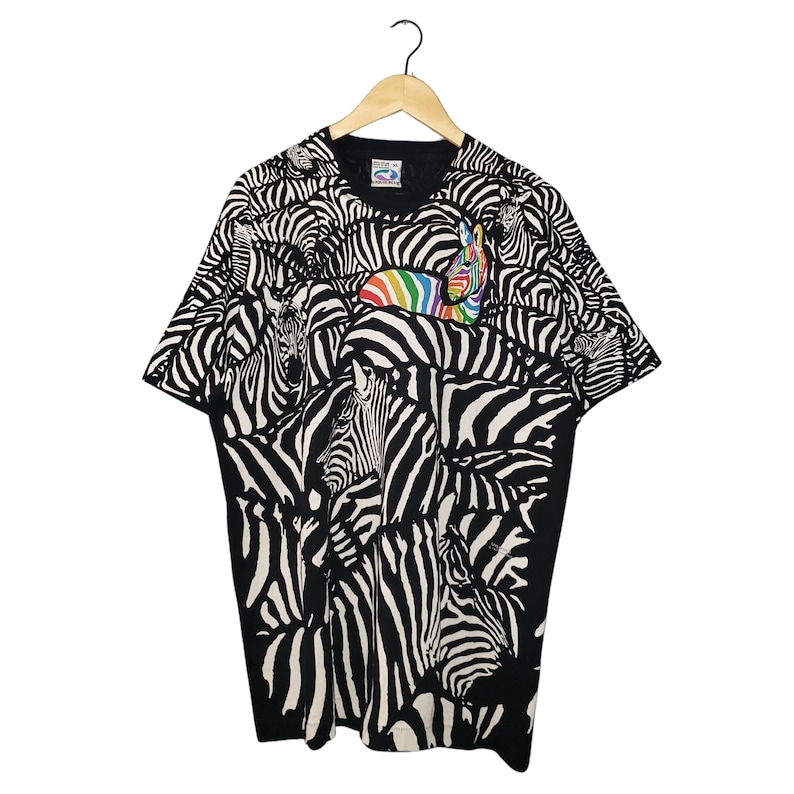 May include: Black and white zebra print t-shirt with a rainbow accent. The short-sleeved shirt features a repeating pattern of zebras. The shirt is hanging on a wooden hanger against a white background.
