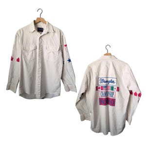 Op de afbeelding: Crèmekleurig Wrangler-shirt met lange mouwen. Rode esdoornbladeren en blauwe sterren sieren de mouwen. De achterkant van het shirt toont het Wrangler-logo en de woorden "Championship Rodeo" met vlaggen.