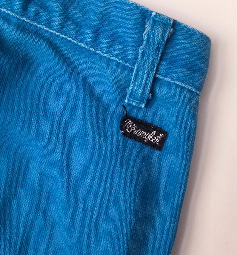 May include: Close-up of a section of bright turquoise denim jeans. The jeans feature a belt loop and a black rectangular tag with the brand name "Wrangler" in white script. The fabric has a textured appearance.