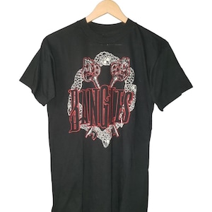 May include: Black t-shirt featuring a graphic design with the word "BANGLES" in red and white lettering. The design includes a decorative border with a snake-like pattern. The shirt is hanging on a wooden hanger.