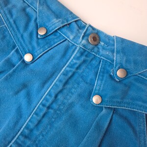 May include: Close-up of the waistband of a pair of bright blue denim jeans. The jeans feature silver metal button closures and decorative stitching. The fabric has a slightly faded appearance.