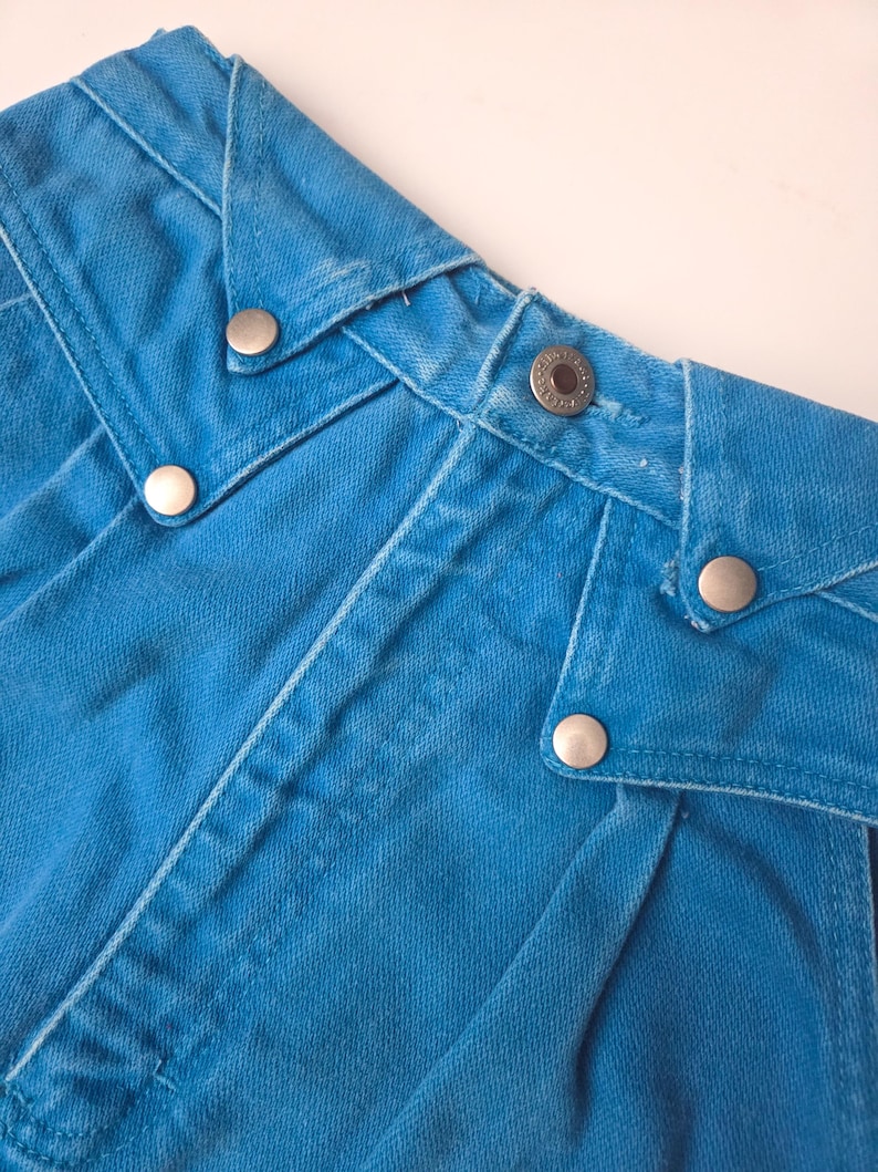 May include: Close-up of the waistband of a pair of bright blue denim jeans. The jeans feature silver metal button closures and decorative stitching. The fabric has a slightly faded appearance.