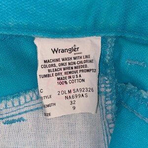 May include: A white care label with black text for a pair of turquoise Wrangler jeans. The label includes the brand name, care instructions, size, style, and length. The label reads: "Wrangler Brand, Machine wash with like colours, only non-chlorine bleach when needed. Tumble dry, remove promptly. Made in USA. 100% Cotton. 2DLM SA92326 NA699AS 32 9"