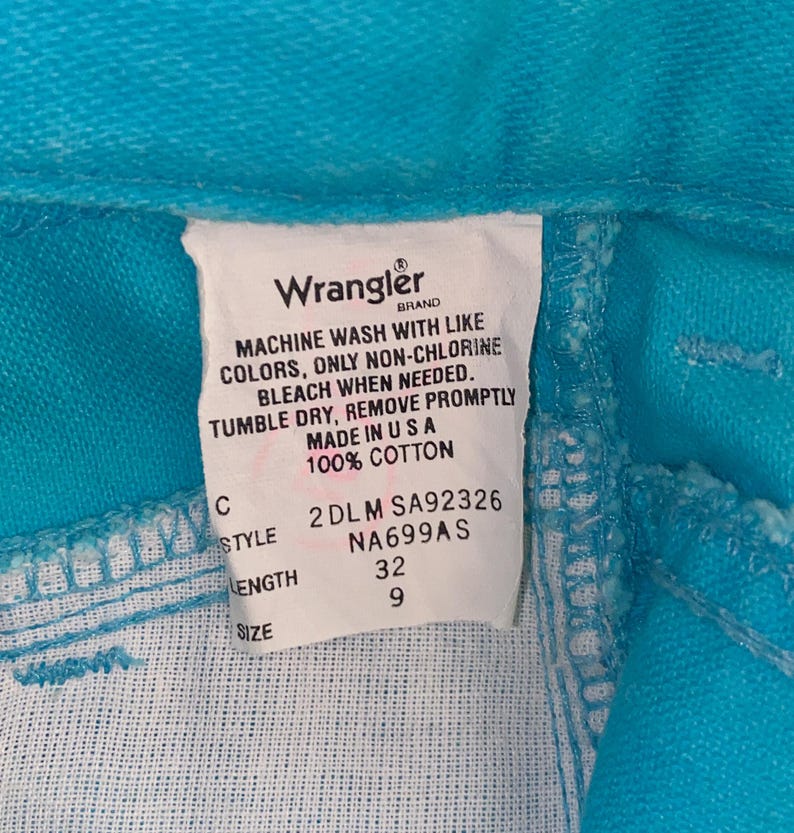 May include: A white care label with black text for a pair of turquoise Wrangler jeans. The label includes the brand name, care instructions, size, style, and length. The label reads: "Wrangler Brand, Machine wash with like colours, only non-chlorine bleach when needed. Tumble dry, remove promptly. Made in USA. 100% Cotton. 2DLM SA92326 NA699AS 32 9"