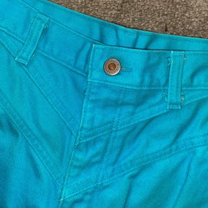 May include: Close-up of the waistband of a pair of turquoise blue jeans. The jeans have a button closure and a single belt loop.