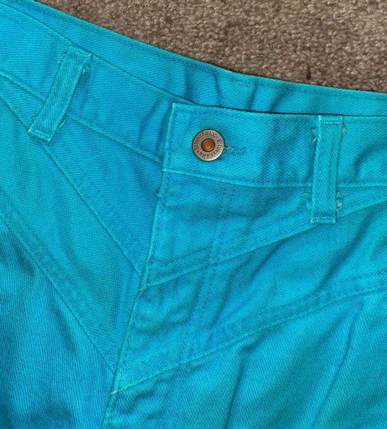 May include: Close-up of the waistband of a pair of turquoise blue jeans. The jeans have a button closure and a single belt loop.