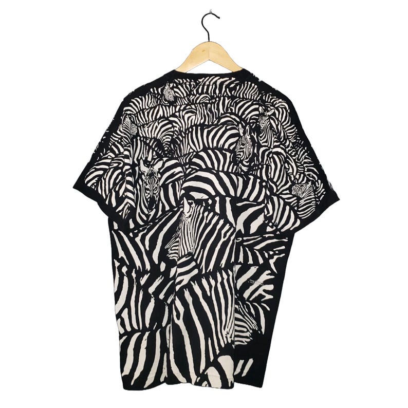 May include: Black and white zebra print t-shirt. The short-sleeved shirt features a repeating pattern of zebra stripes, creating a bold and eye-catching design. The shirt is hanging on a wooden hanger against a white background.