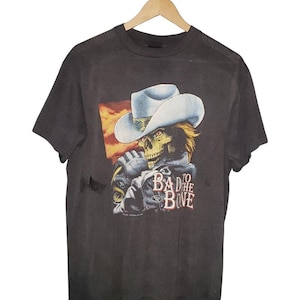 Może przedstawiać: Czarny vintageowy t-shirt z grafiką przedstawiającą szkielet w kowbojskim kapeluszu i skórzanej kurtce. Pod obrazkiem znajduje się napis "To the Bad to the Bone".