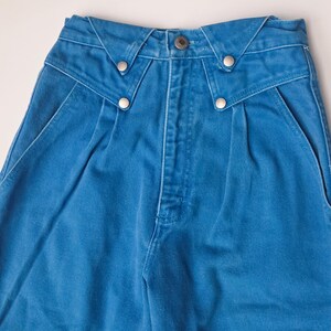 May include: Close-up of blue denim pants. The pants have a high waist with a unique design at the waistband, featuring silver-colored button details. The front of the pants has pleats and pockets.