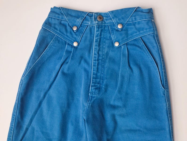 May include: Close-up of blue denim pants. The pants have a high waist with a unique design at the waistband, featuring silver-colored button details. The front of the pants has pleats and pockets.